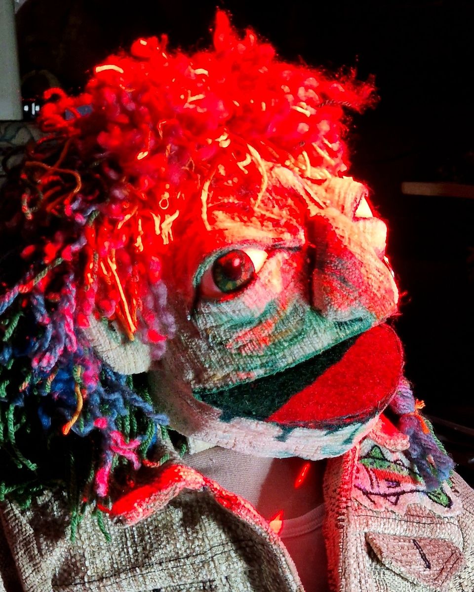 GOOBIE, a finished puppet with yarn hair and an upcycled jacket under red stage light